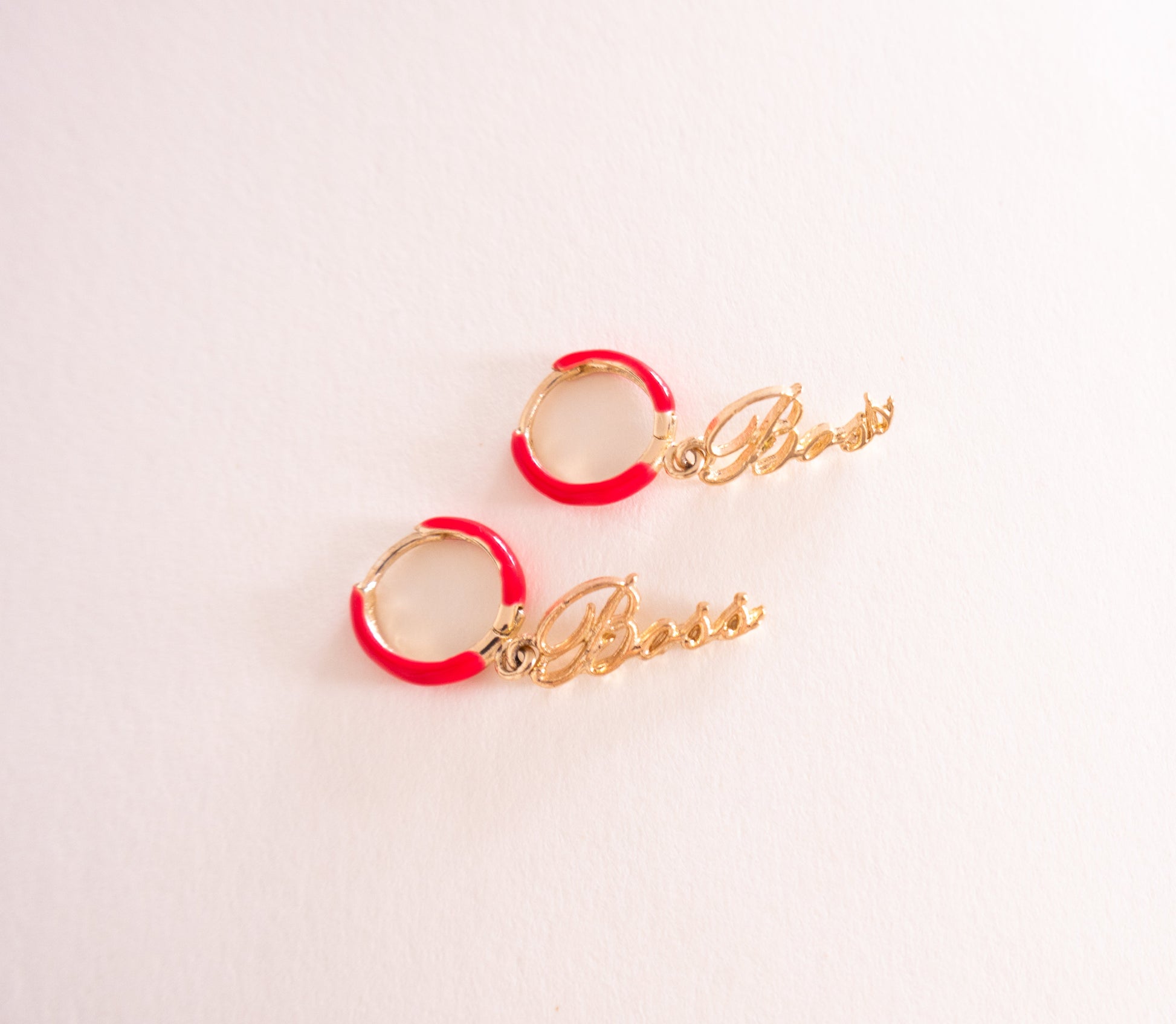 Pair of earrings with red loops and gold 'Boss' charms on a light pink background