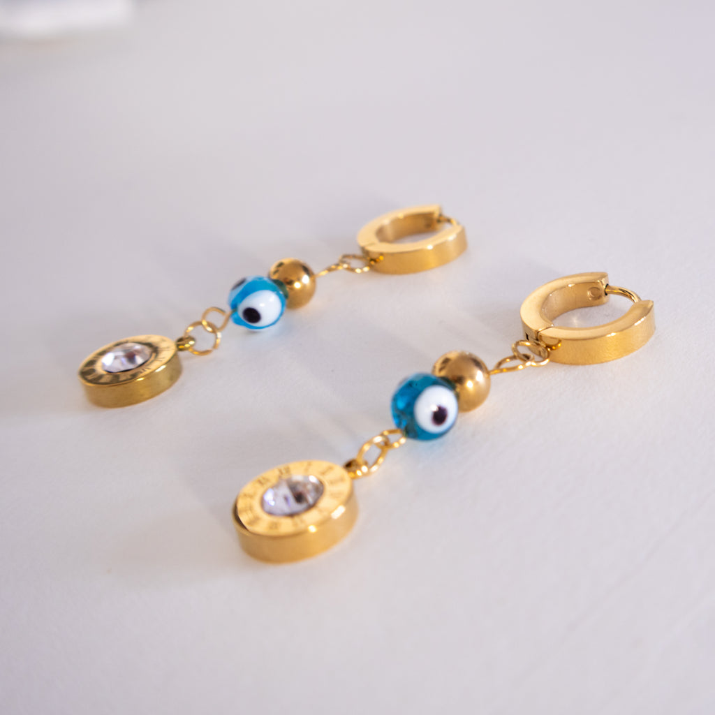 Gold hoop earrings with blue evil eye charms on a light gray background