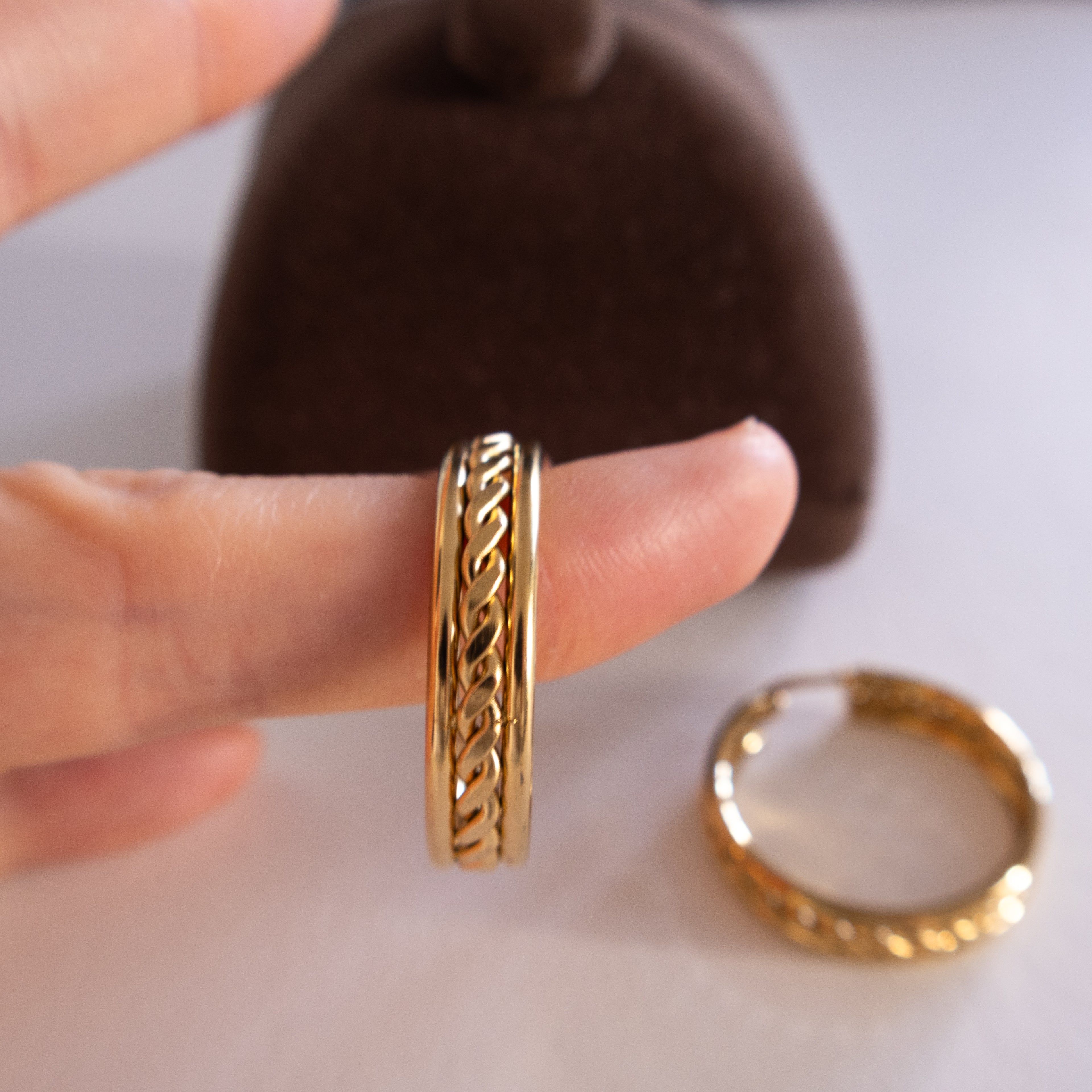Gold ring with intricate design worn on a finger, with another ring on a white surface.