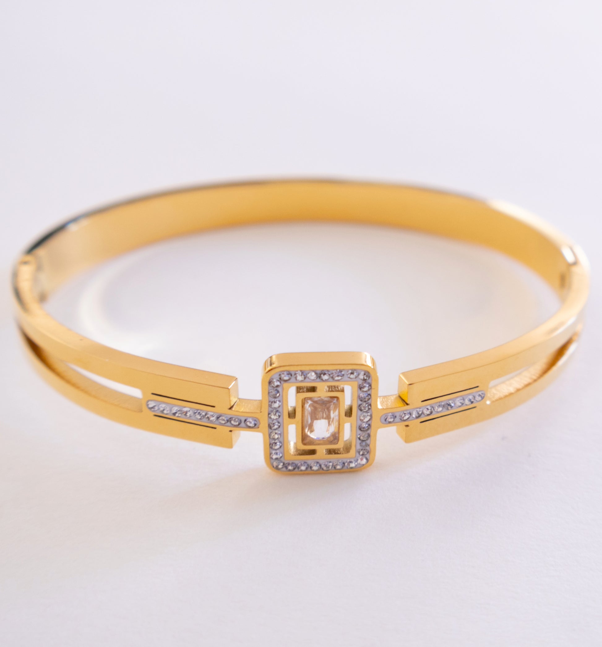 Gold bracelet with a square design and clear stones on a light gray background