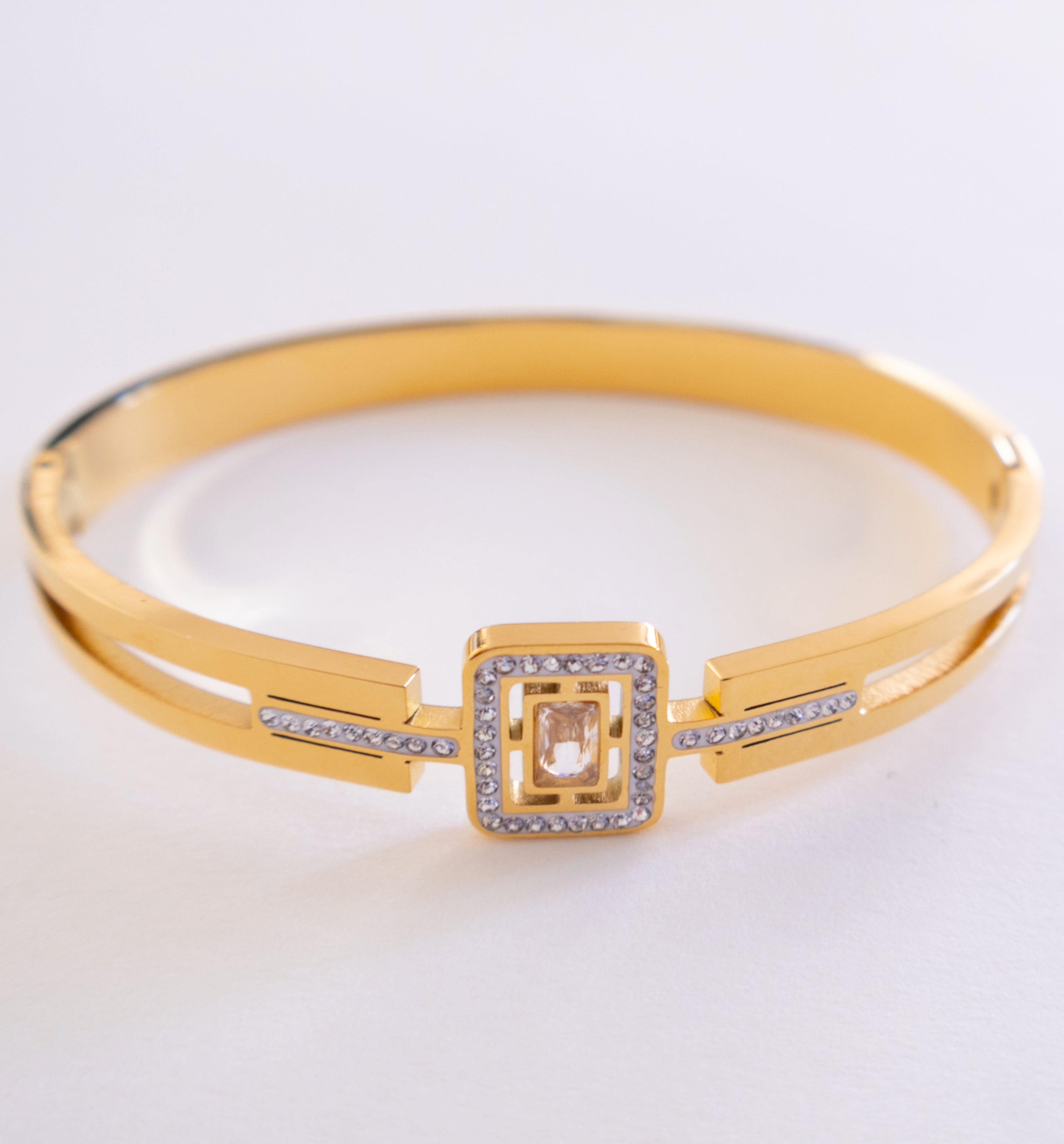 Gold bracelet with a square design and clear stones on a light gray background
