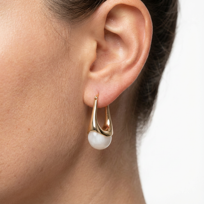 Gold earring with a pearl drop worn by a person on a neutral background
