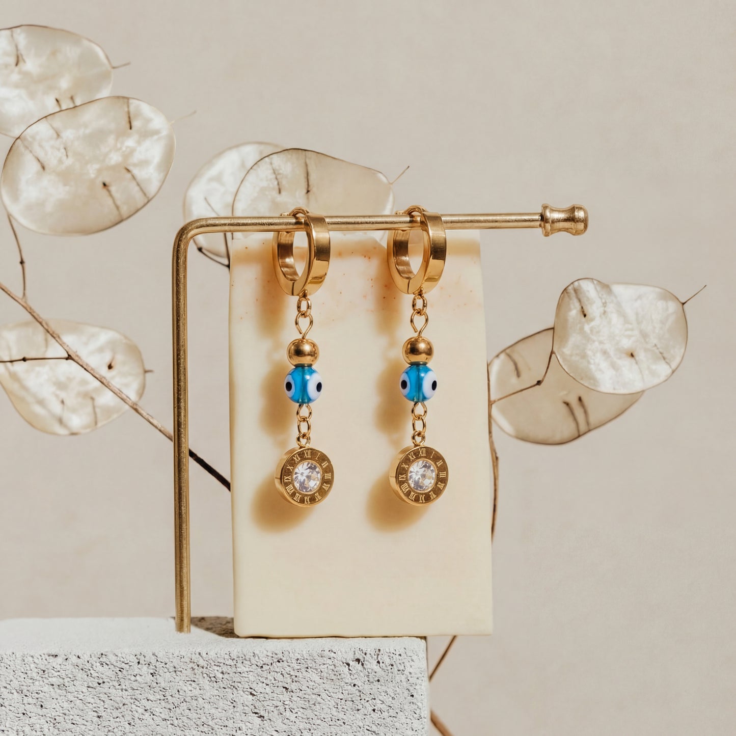 Gold earrings with blue and clear stones on a gold stand against a beige background with decorative leaves.