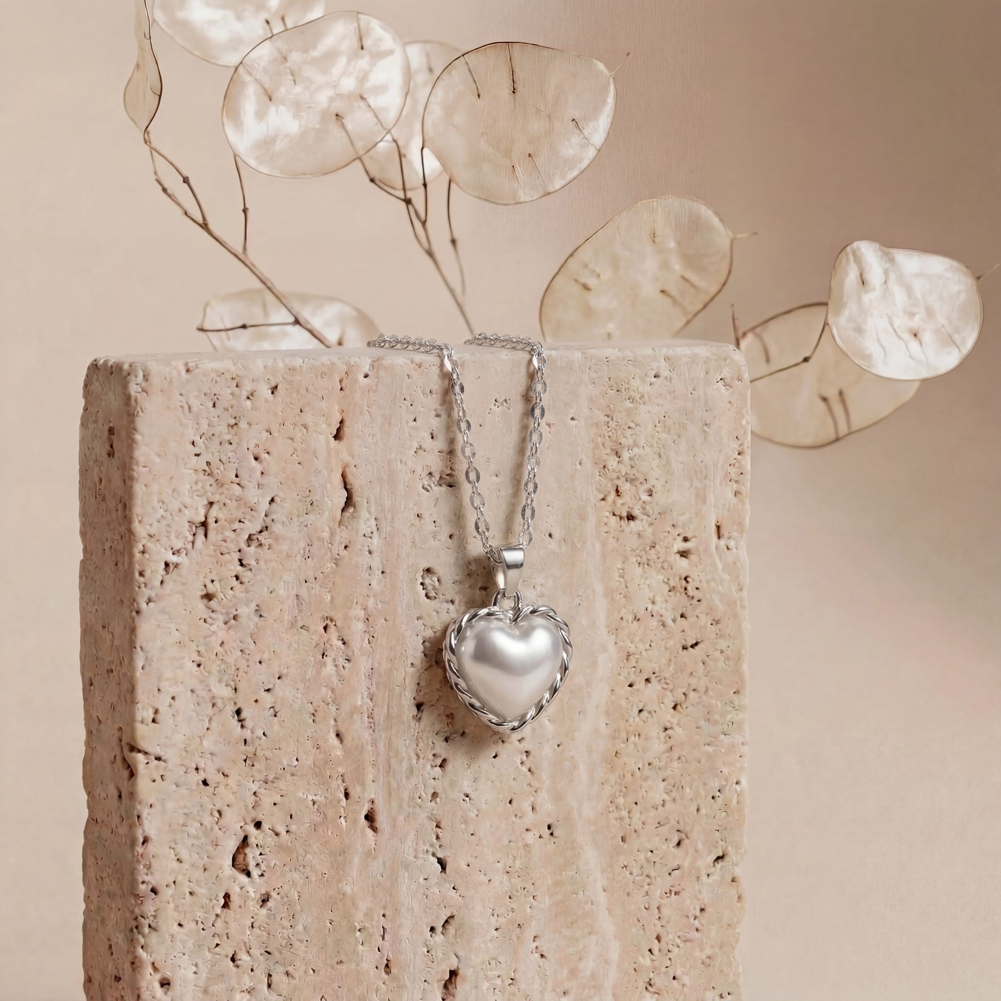 Heart-shaped silver pendant on a chain against a textured stone surface with decorative leaves.