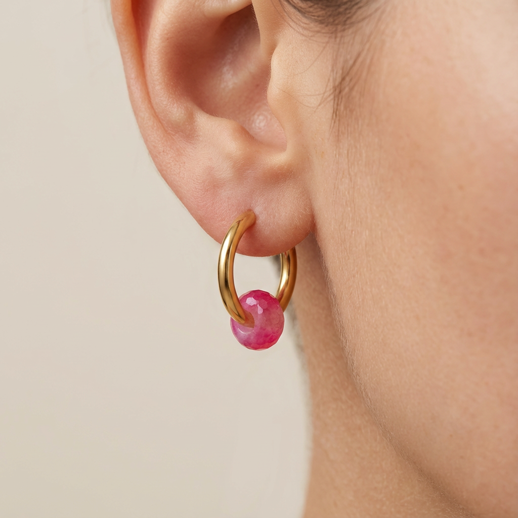 Gold hoop earring with a pink gemstone worn by a person on a beige background