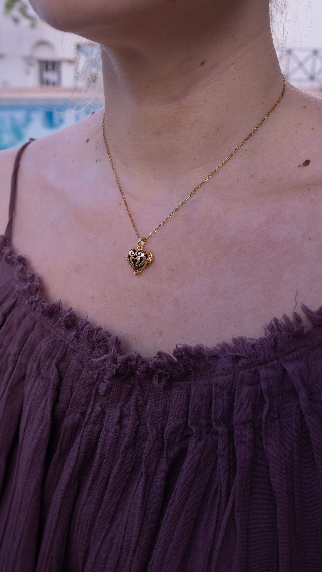 Gold necklace with a heart pendant worn by a person in a purple top, with a blurred background.