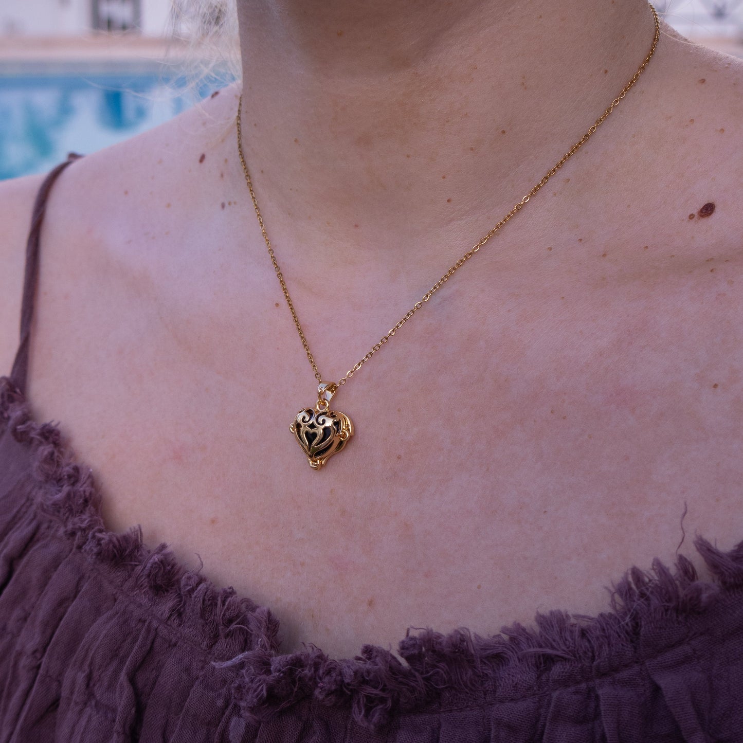 Gold necklace with a heart pendant worn by a person in a purple top, with a blurred background.