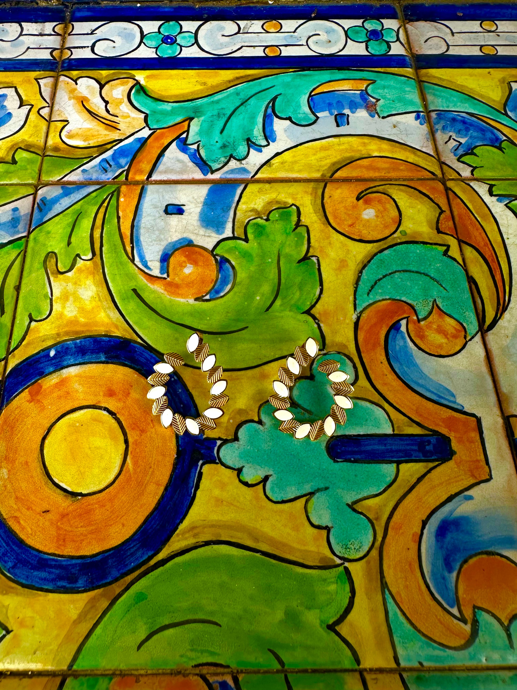 A close-up of a colorful hand-painted ceramic tile with abstract green, yellow, blue, and orange scrollwork, featuring two small white floral details earings.